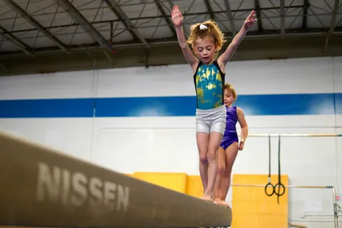 A young gymnast balancing on a beam with her arms raised while another waits her turn